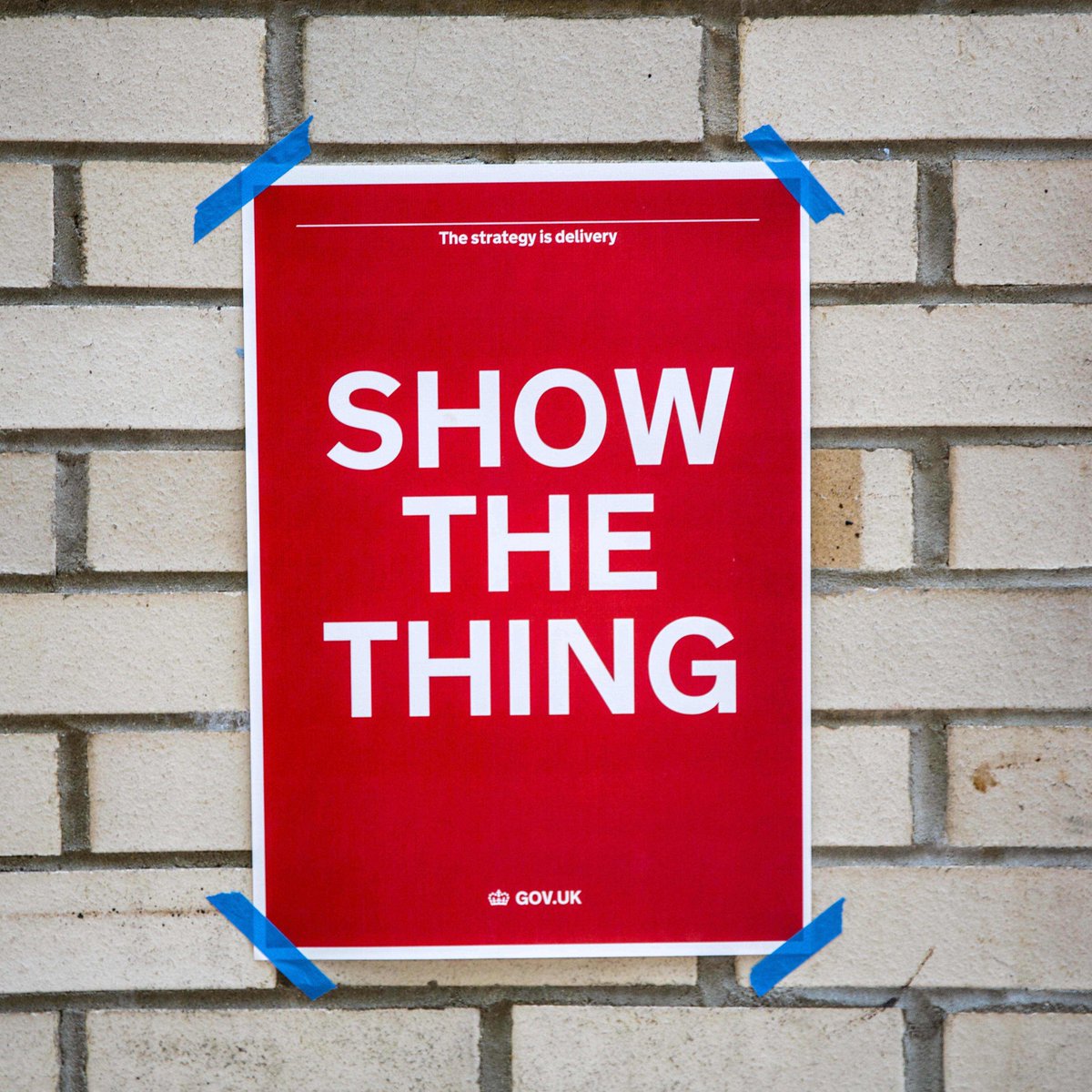 A poster taped to a wall with the words 'show the thing' prominent in the center and the words 'The strategy is delivery' in small font above and 'gov.uk' below